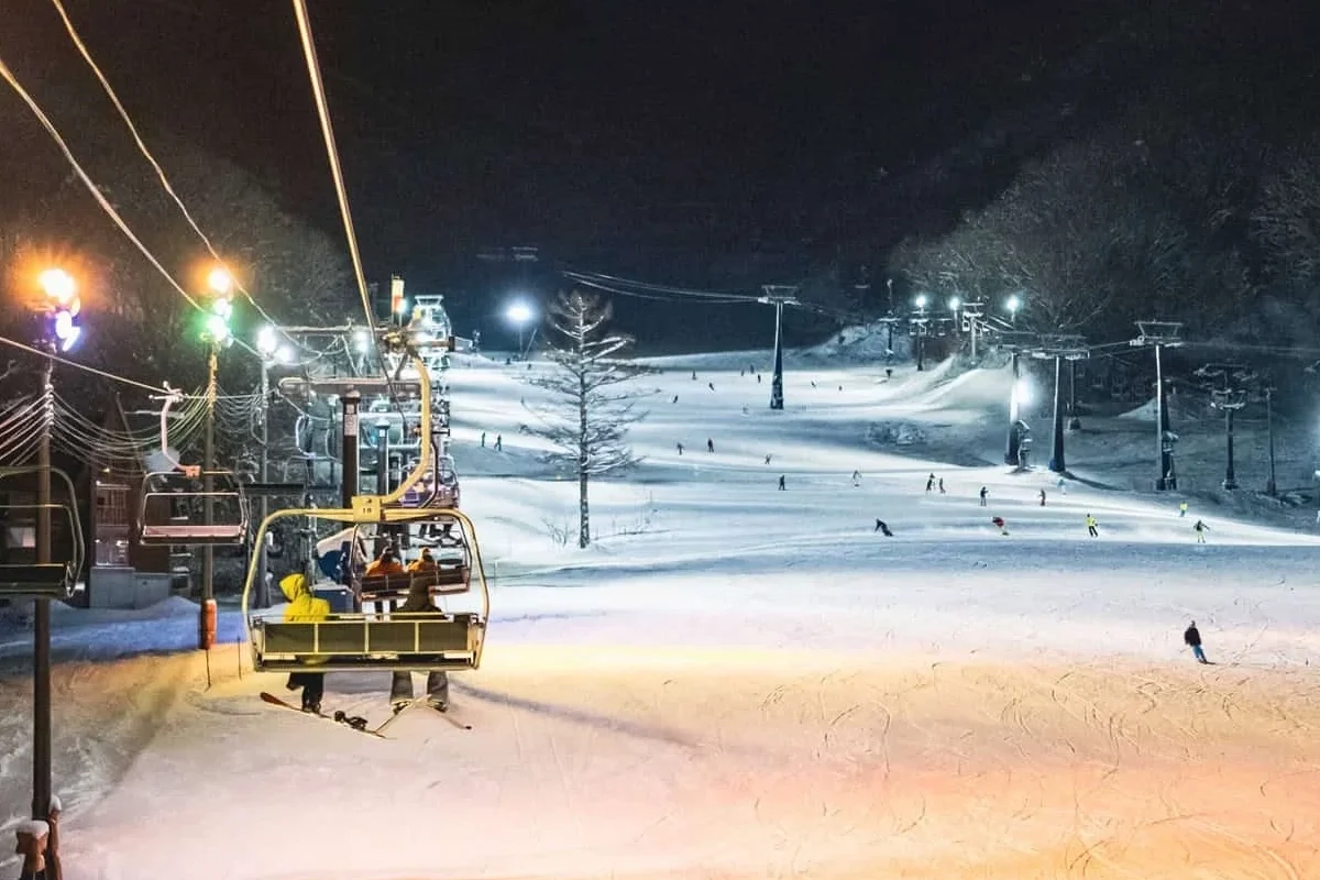 Chairlifts and skiers on a brightly lit ski slope during a night skiing session at Hakuba Goryu, Japan.