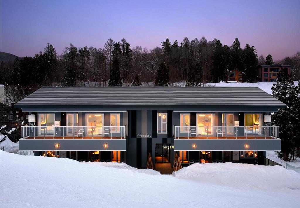Modern two-story ski lodge with glowing windows surrounded by deep snow and pine trees at dusk.
