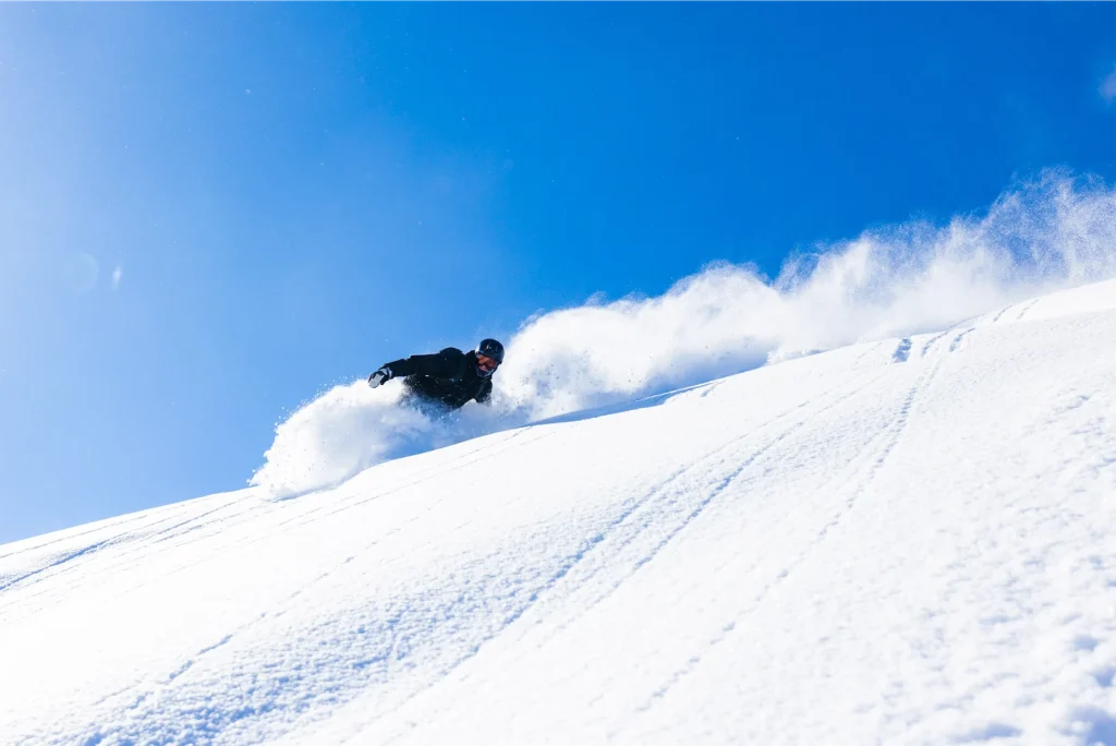 Snowboarder carving through deep powder snow on a steep sunny slope under a clear blue sky.