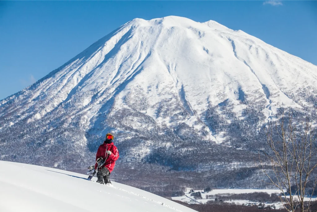 Snowboarder in red jacket holding a board on a snowy peak with snow-capped Mount Yotei in the background.