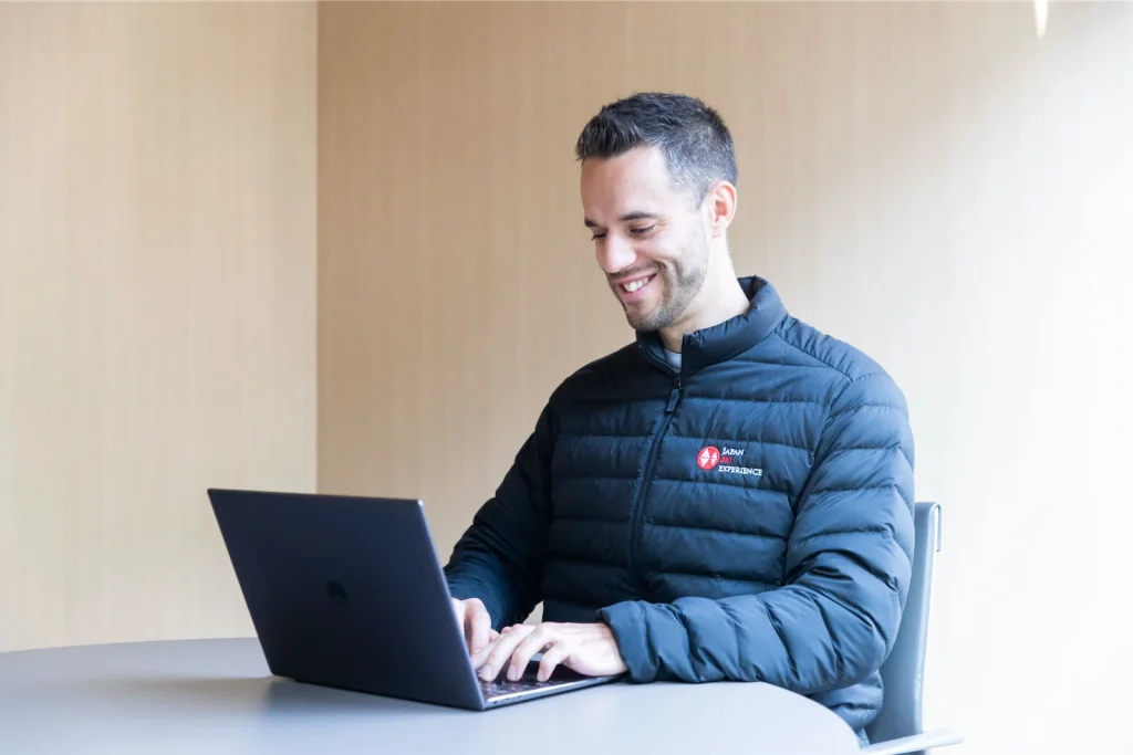 Smiling man wearing a dark jacket typing on a laptop at a desk indoors.