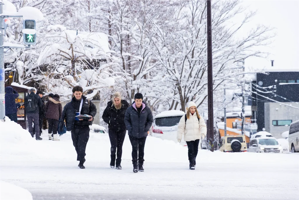 Pedestrians in winter coats walking across a snow-covered town street in a ski resort village.