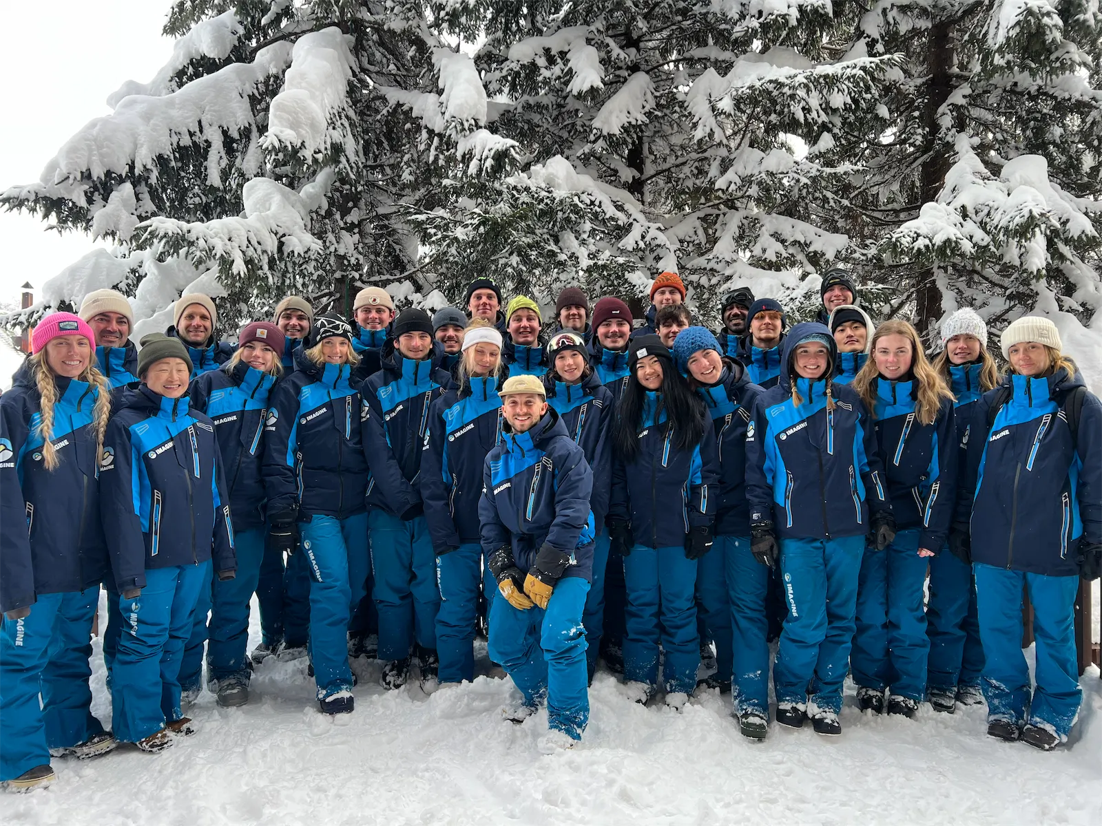 Large team of ski instructors wearing matching blue winter uniforms posing together outdoors in front of snow-draped trees.