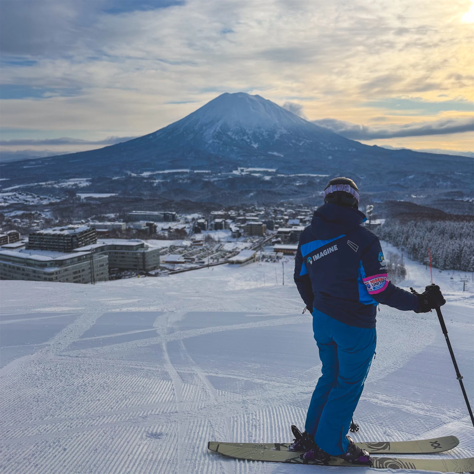 Skier standing on a groomed snow run looking out toward Mount Yotei and the resort village below.