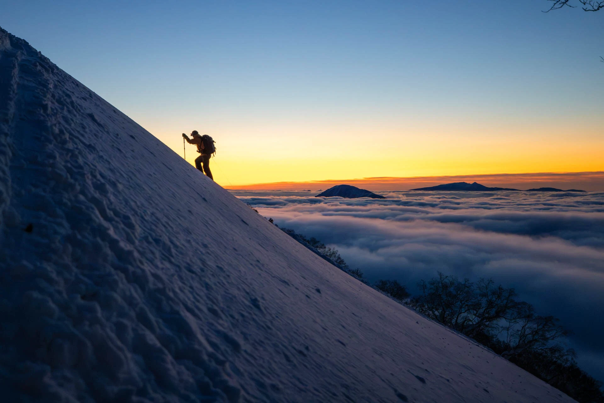 Silhouette of a backcountry skier ascending a steep snow slope at sunrise above a sea of clouds.