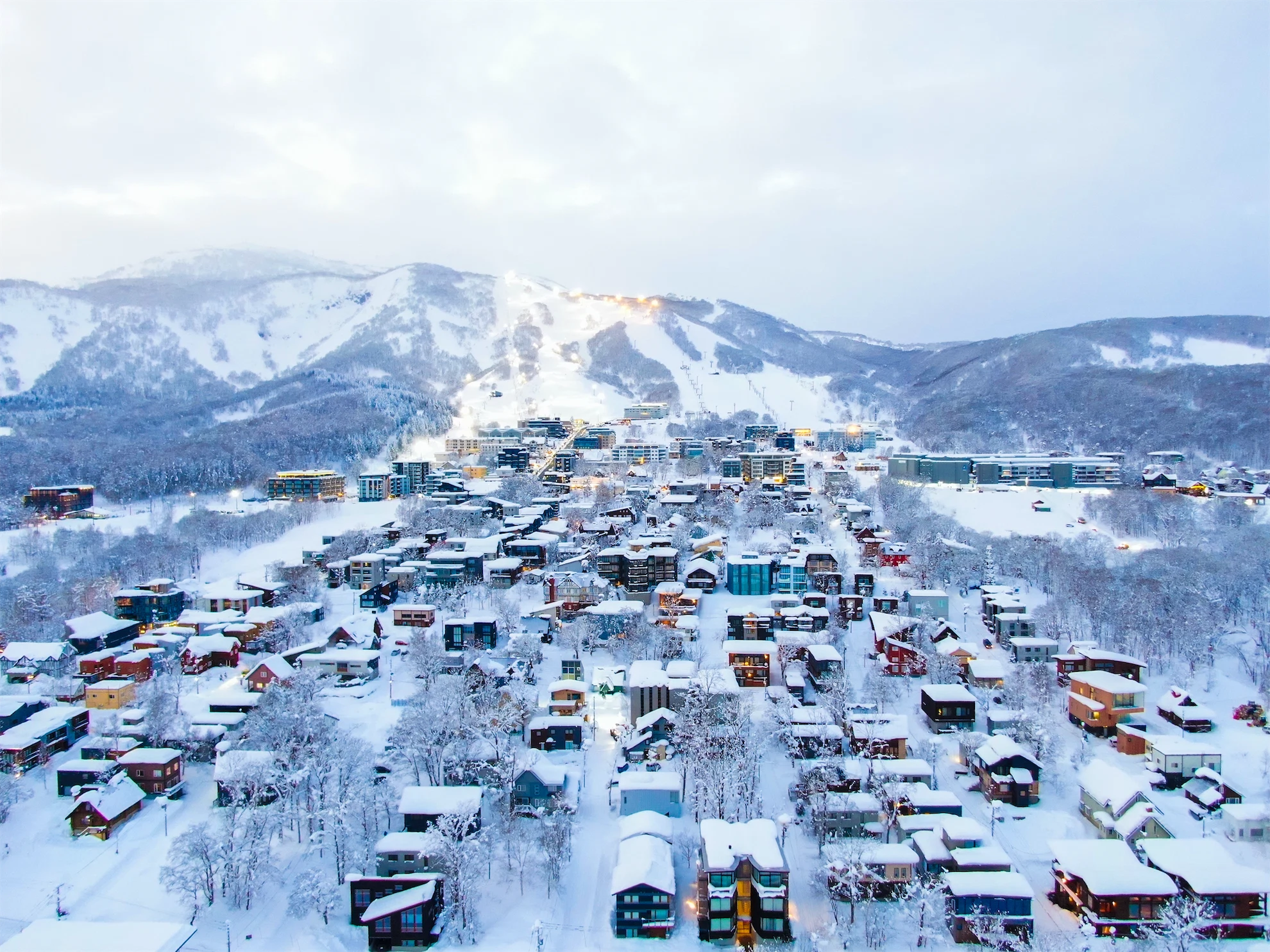Aerial view of snow-covered Hirafu village at twilight with lit-up ski slopes in the background.