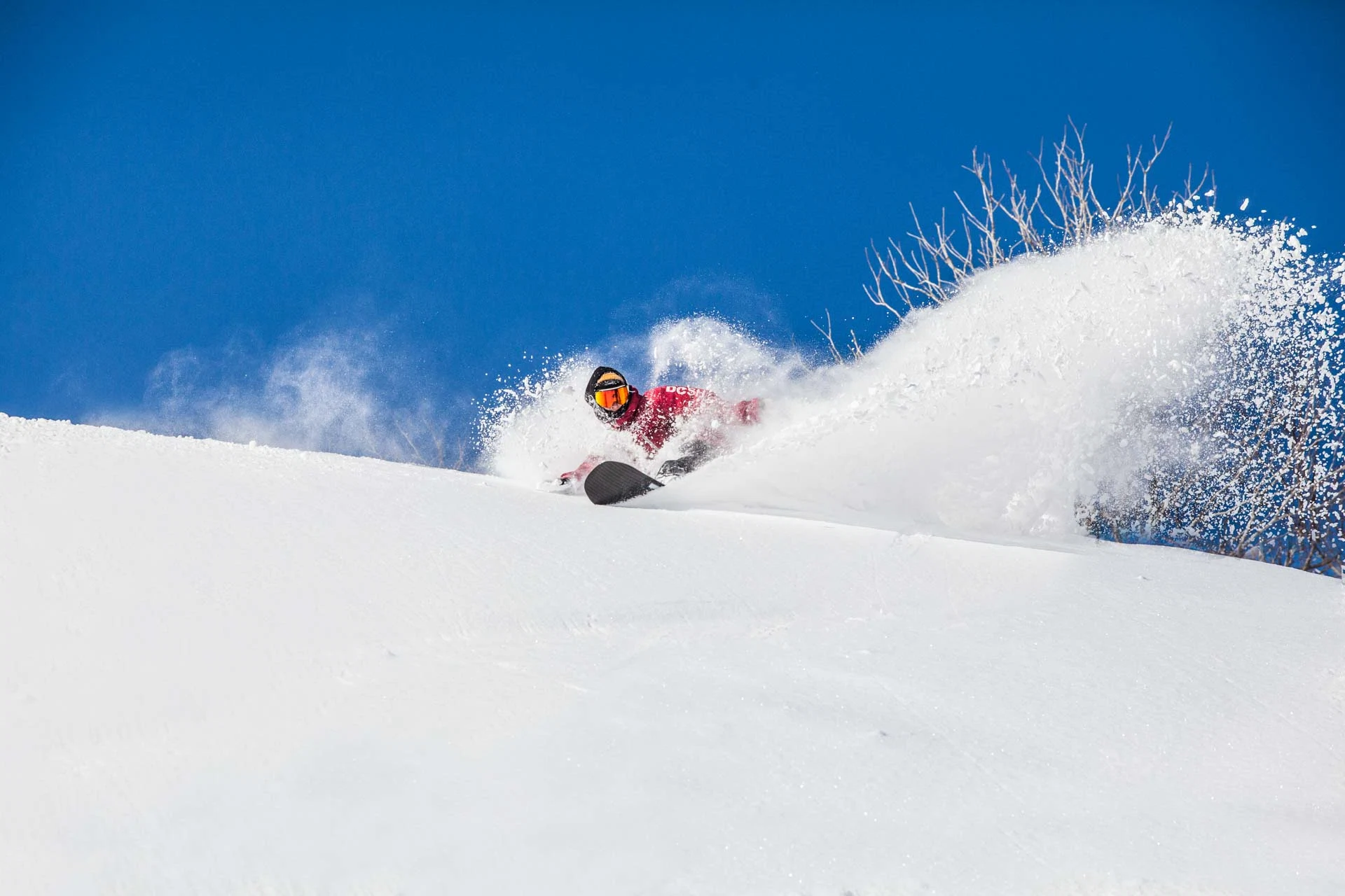 Snowboarder in a red jacket carving a sharp, high-spray turn against a clear blue sky.