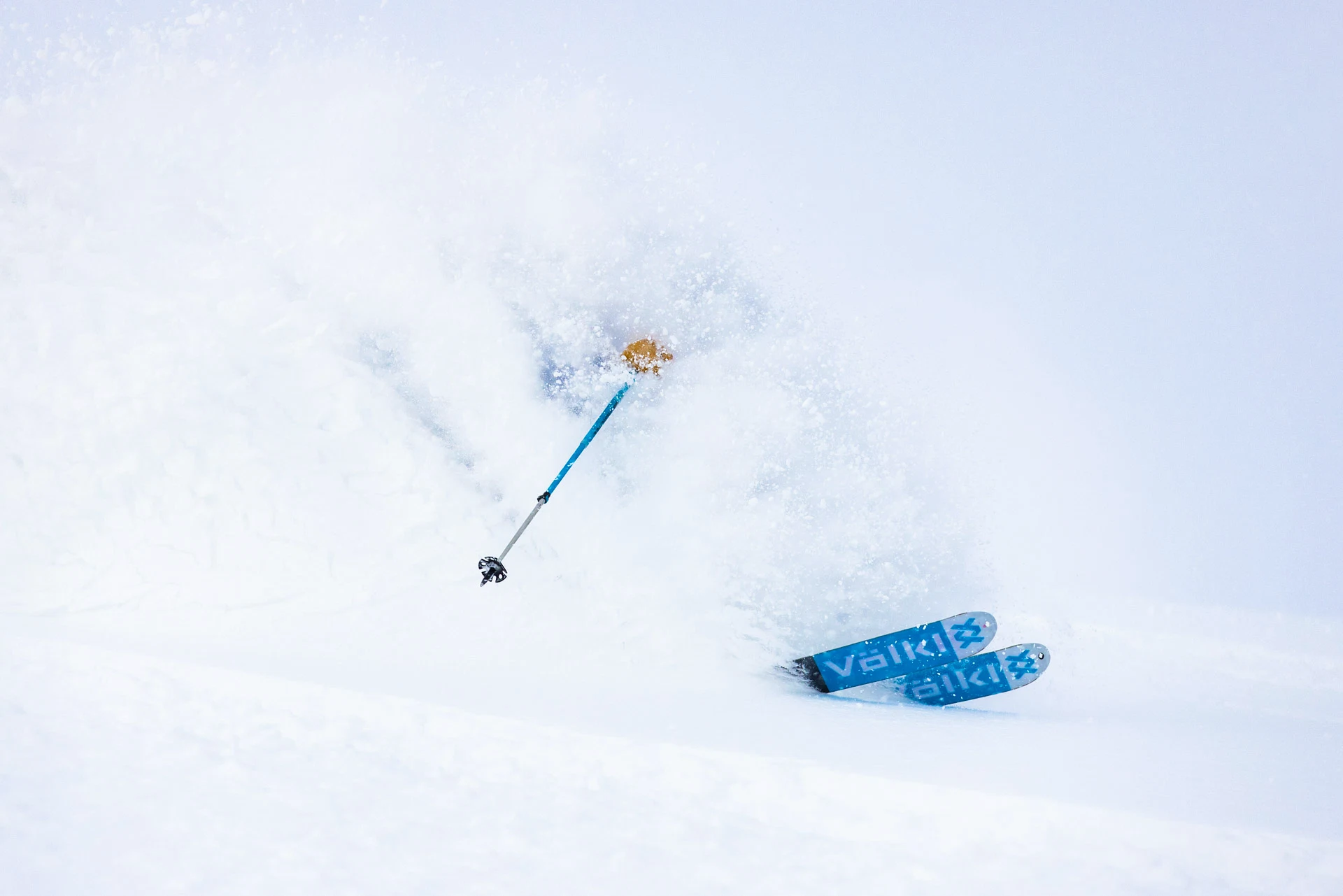 Skier nearly submerged in deep powder snow with Völkl skis carving a turn.