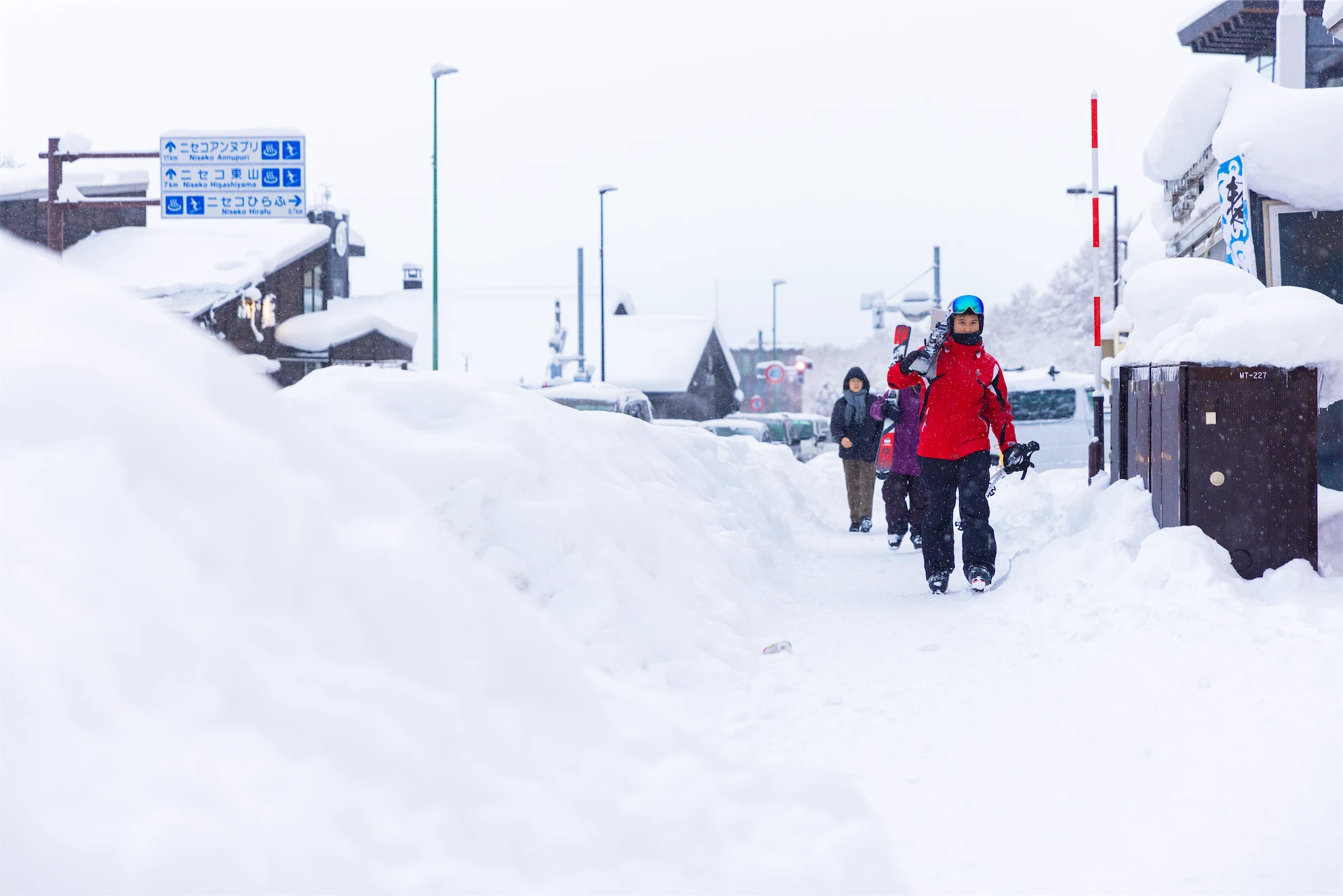 Person carrying skis walking along a heavily snow-covered town sidewalk next to high snowbanks.