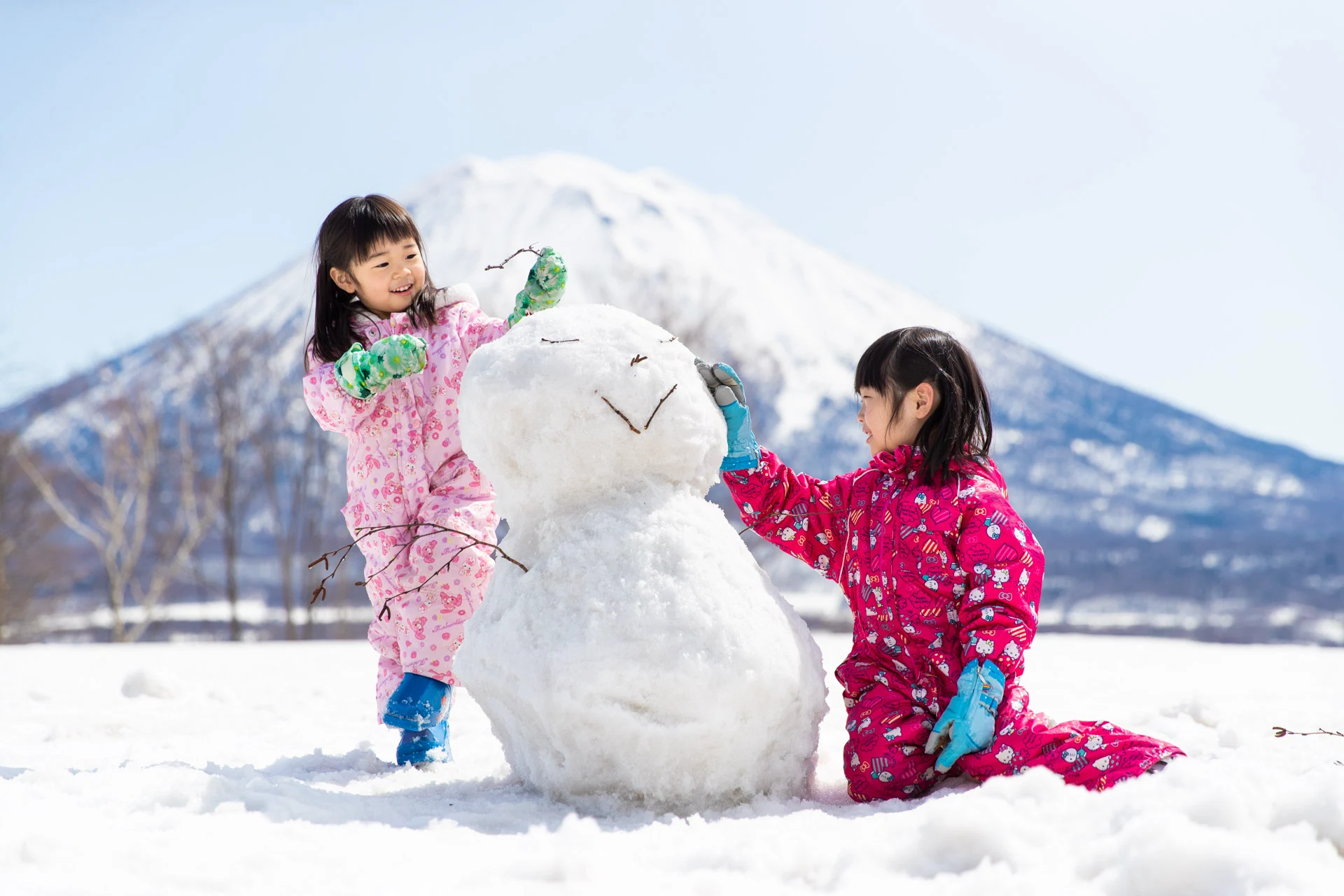 Two young children in pink snowsuits building a snowman with Mount Yotei in the background.