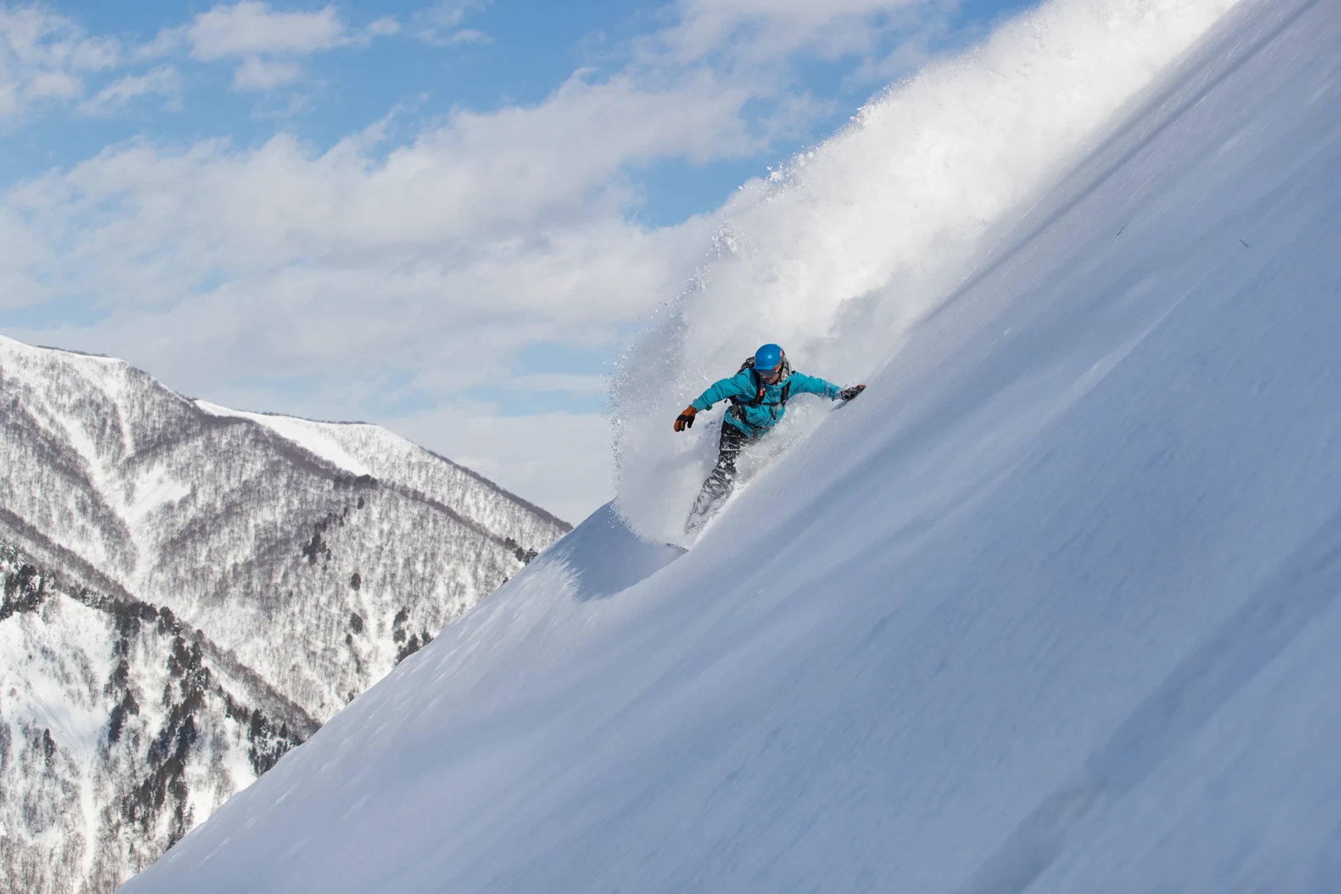 Snowboarder in a bright blue jacket carving down a steep powder slope under a blue sky.