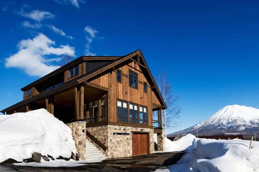 Exterior of a multi-story timber and stone chalet set against deep snow with Mount Yotei in the background under a clear sky.